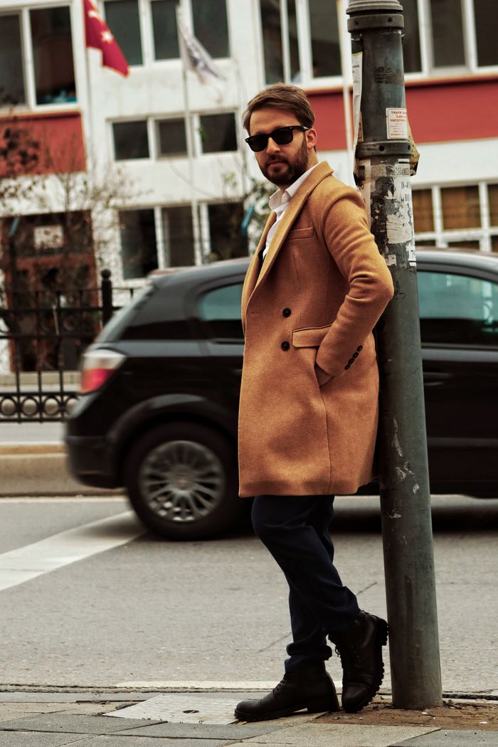 Fashionable man in a brown coat and sunglasses leaning on a post in a city street setting.