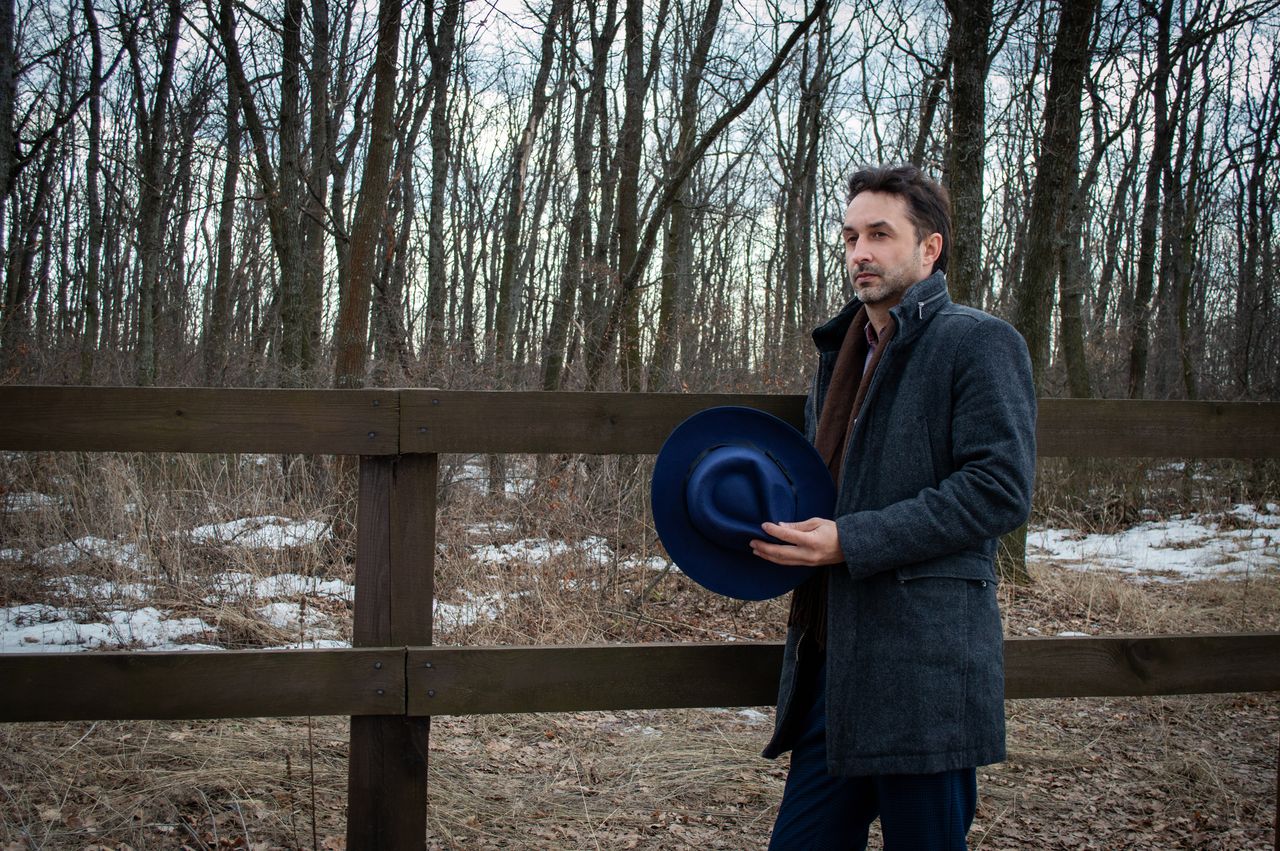 Thoughtful adult bearded male in warm casual clothing holding hat and looking away while standing next to wooden construction on cloudy day