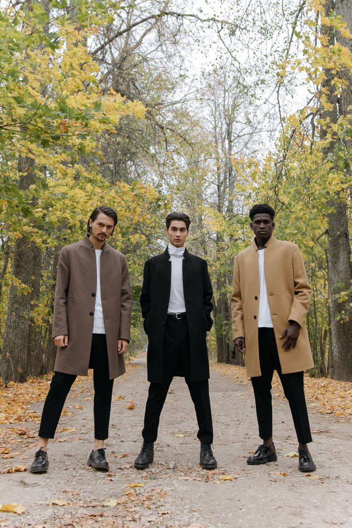 Three stylish men in coats walking through a tree-lined autumn park pathway filled with fallen leaves.